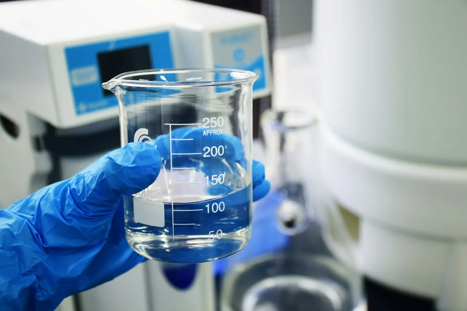 A gloved hand holding a chemical beaker with clear liquid in a laboratory