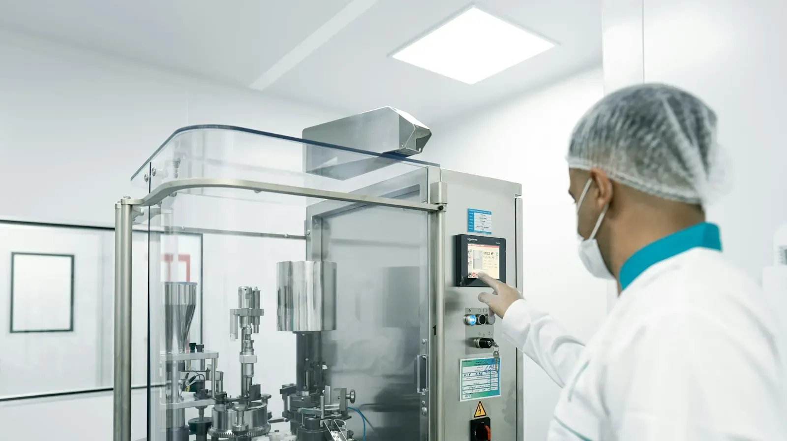 A worker in a white coat, cap and mask next to industrial equipment at a production site